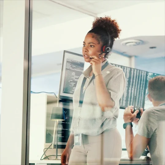 Customer support agent, woman with a headset, speaking on a call in an office setting with computer monitors.