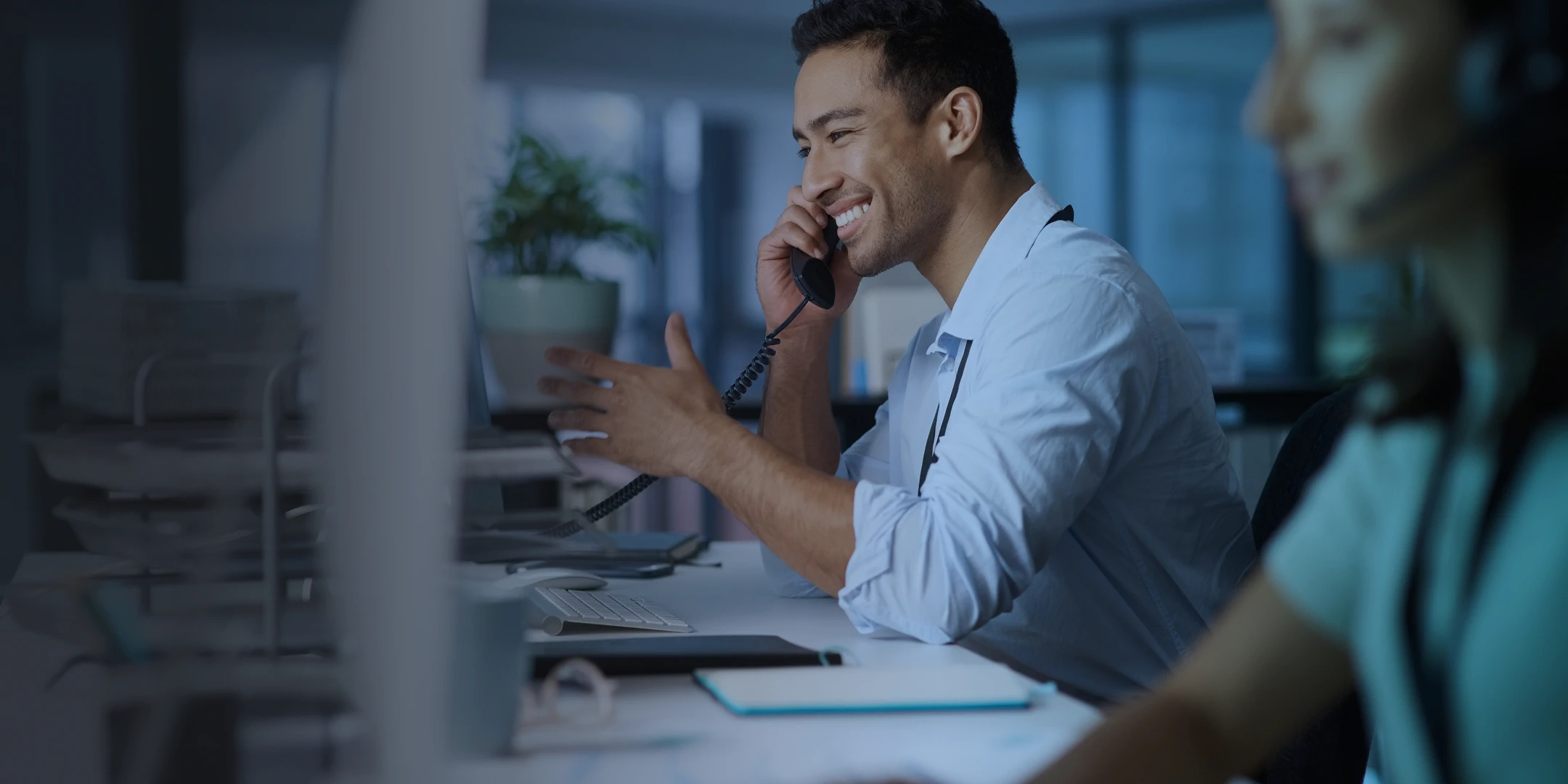 A smiling man in a light blue shirt enthusiastically gestures while talking on a corded phone in a modern office setting.