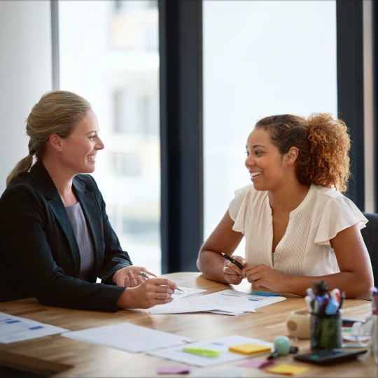Two smiling businesswomen strategizing sales and success at a conference table in an office.