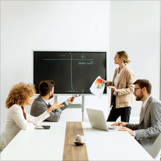 Four businesspeople in a meeting room as a woman presents a pie chart on a large screen with plus-minus chart, while colleagues in suits review laptops and listen.