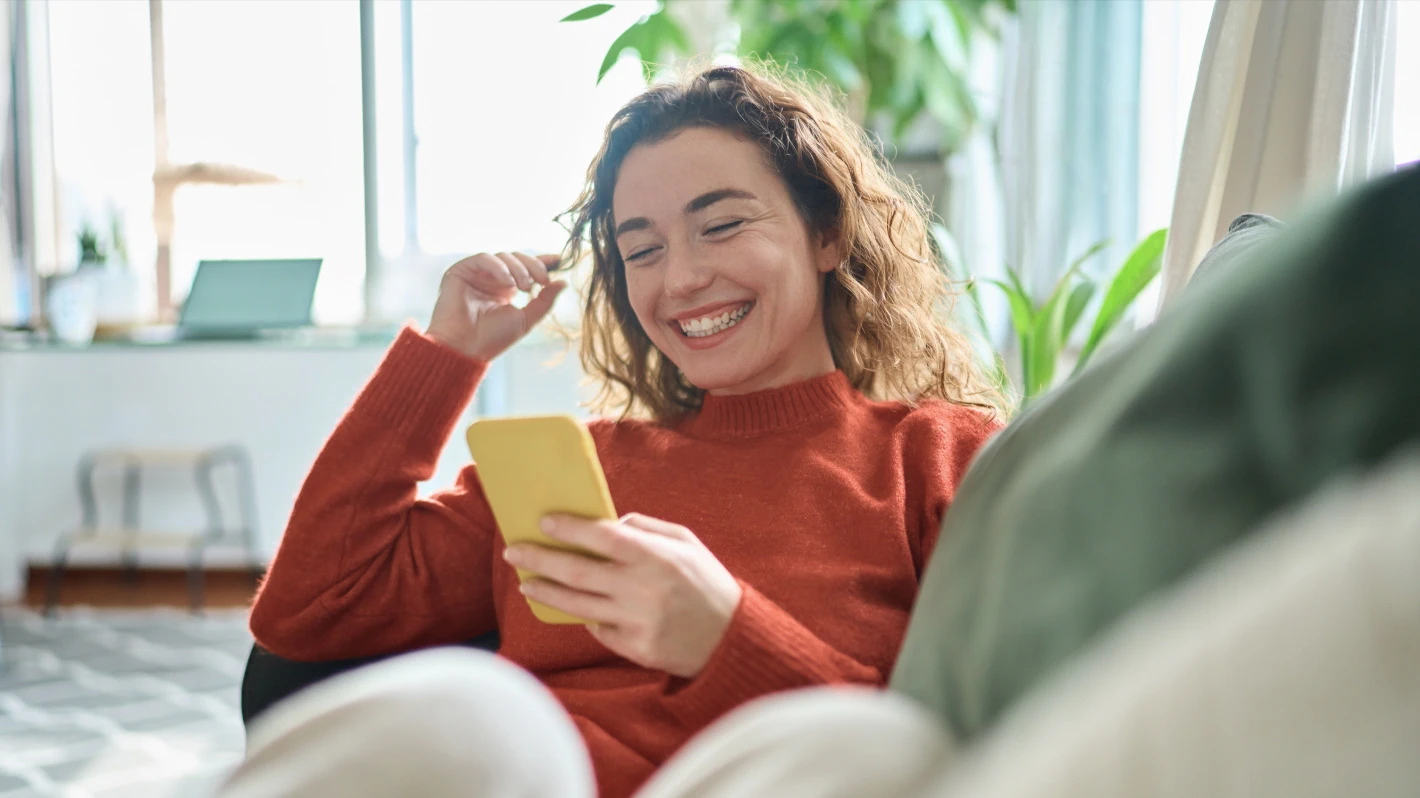 A young, smiling woman with curly hair is relaxing on a couch with plants in the background, looking at her phone