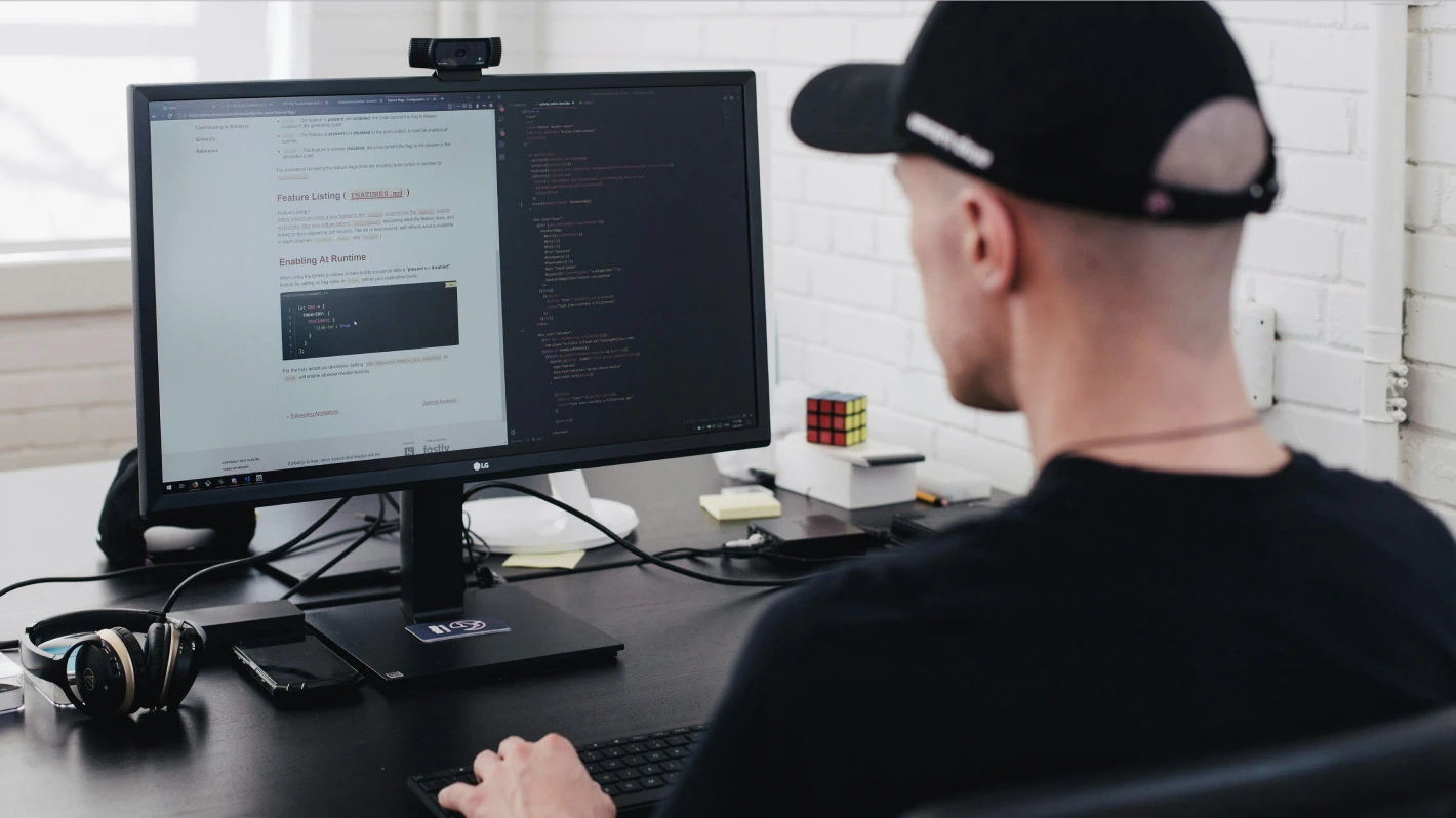 A man wearing a baseball cap is working at a computer in a large, bright office