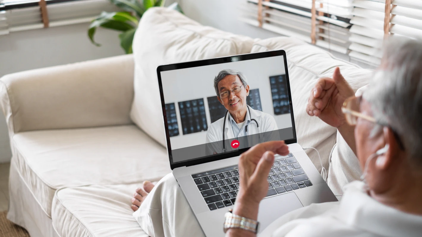 An elderly patient has a video consultation with a doctor on his laptop while sitting on a couch at home