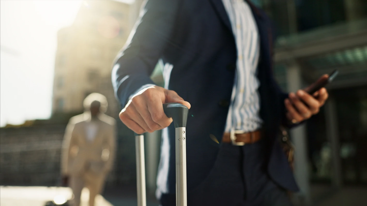 Close-up on a business man wearing a suit and holding the handle of a suitcase and his smartphone