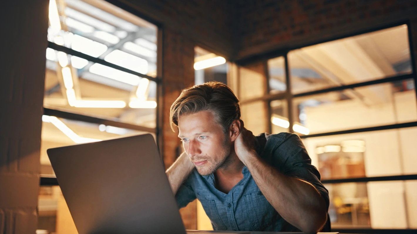 A blond man in a blue shirt is working intently at a laptop, with his hands behind his neck