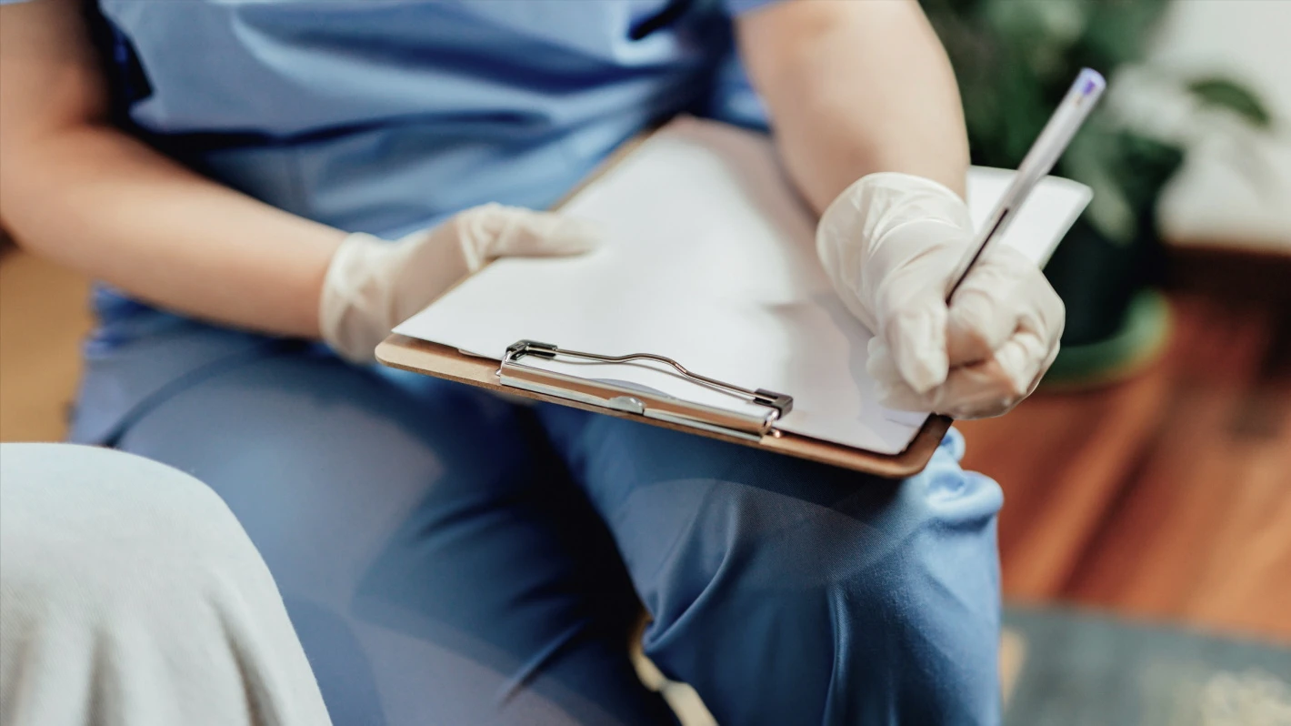 A medical worker wearing blue scrubs and white gloves is holding a clipboard and writing with a pen