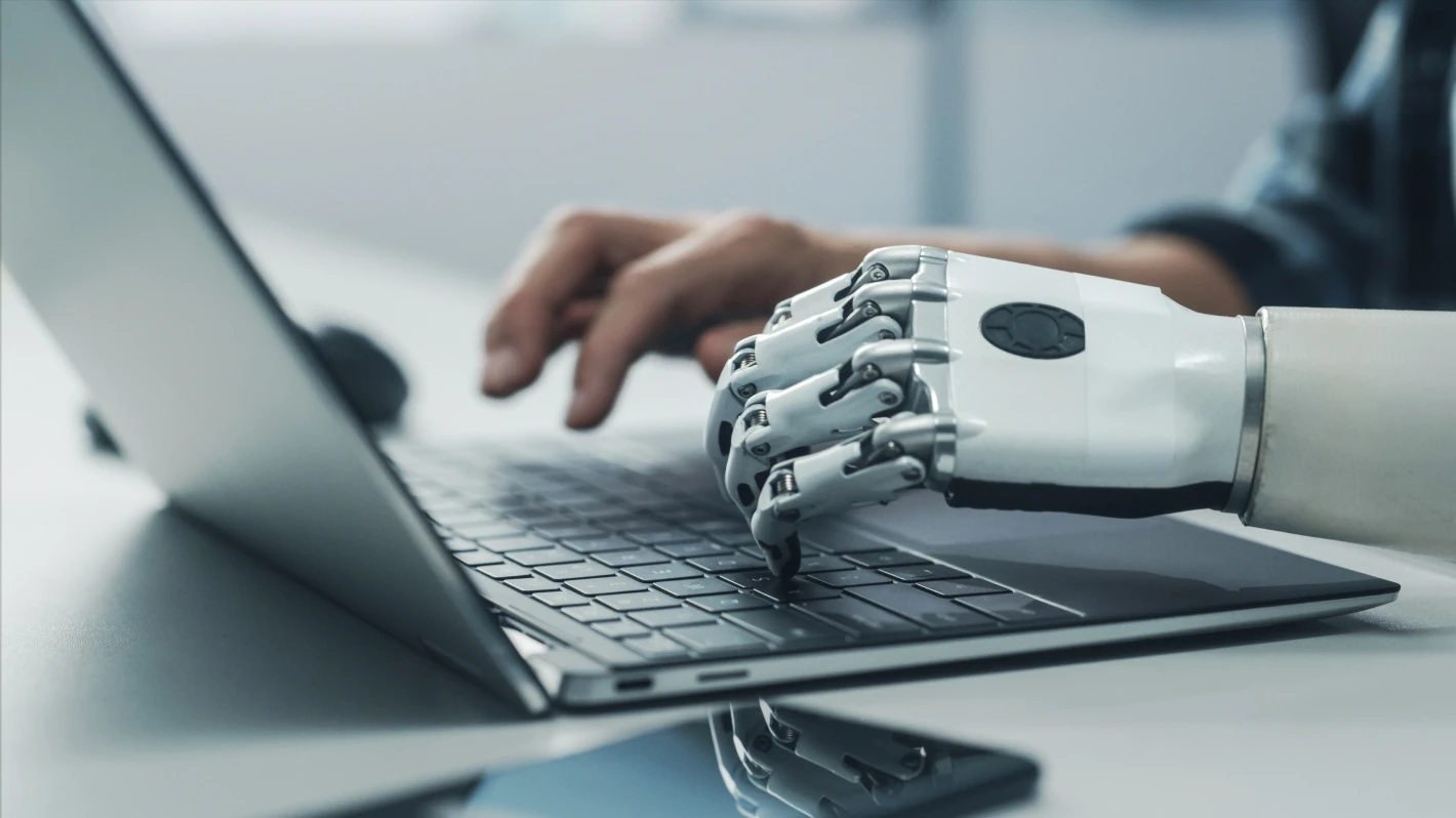 A human hand and a robotic hand are both typing on a laptop keyboard