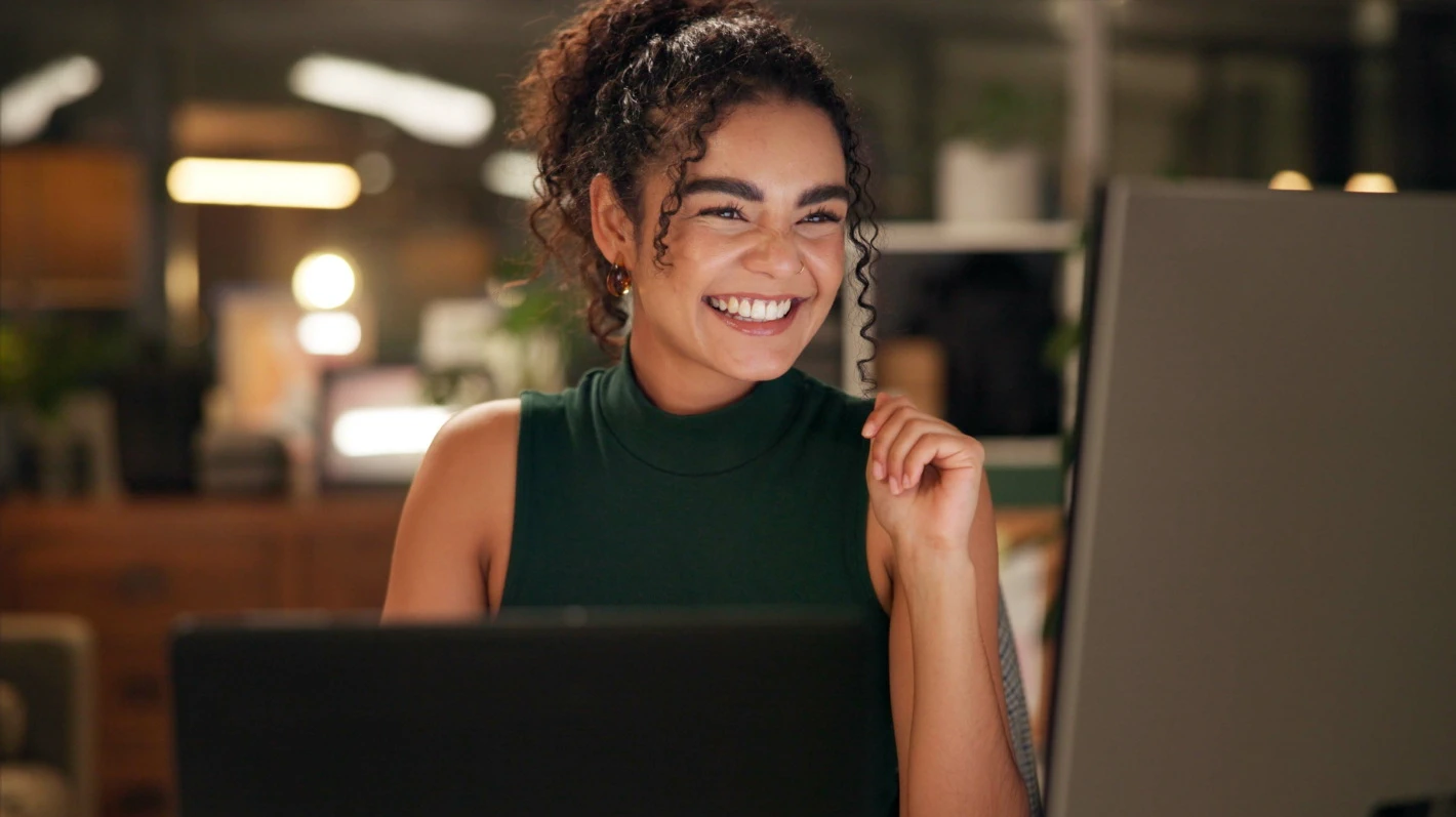 A smiling woman with black curly hair, wearing a sleeveless T-shirt, working on a laptop at a desk