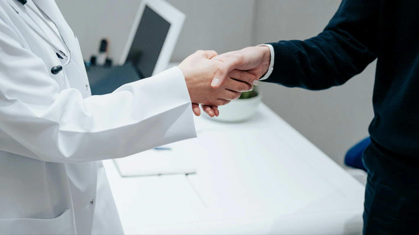 A doctor in a white coat with a stethoscope shakes hands with a patient over a desk in a medical office