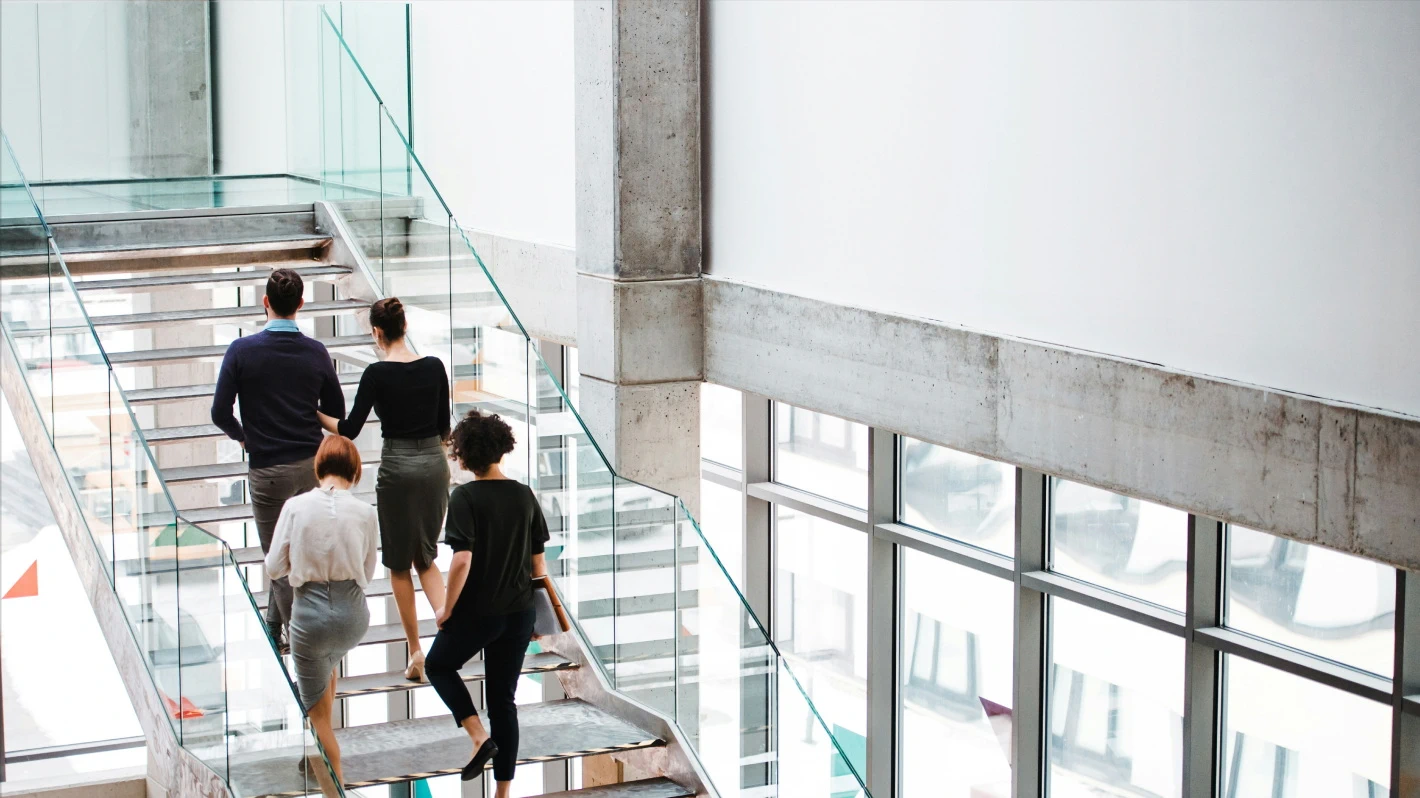 Four people walk upwards on a modern staircase with glass railings in a bright, open building
