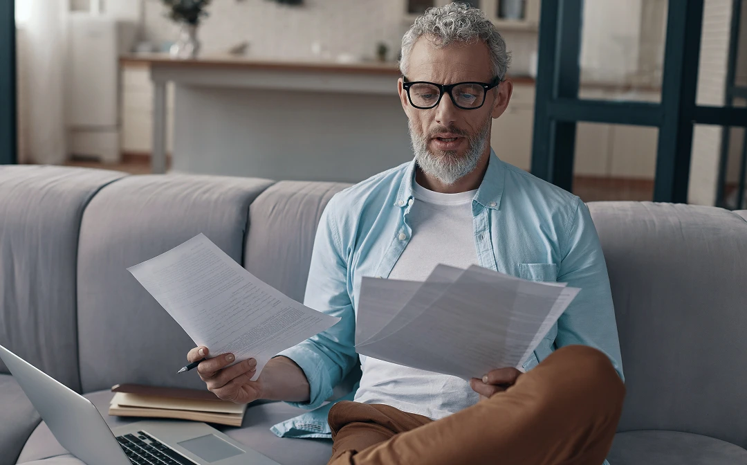 Older man with gray hair and glasses reviewing medical bills on a sofa, wearing a white t-shirt and blue shirt