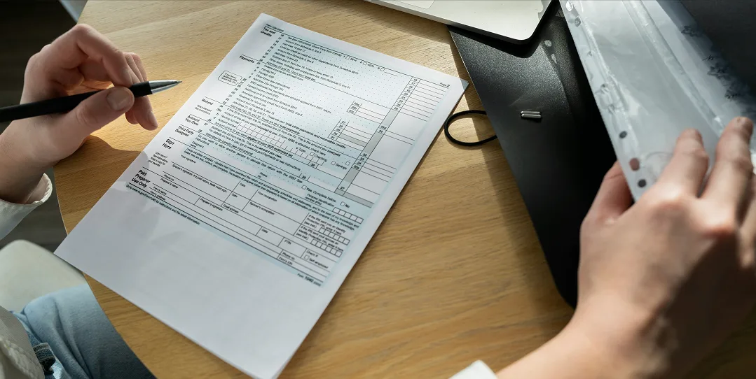 Person holding a pen while filling out a tax form on a light-colored wooden desk