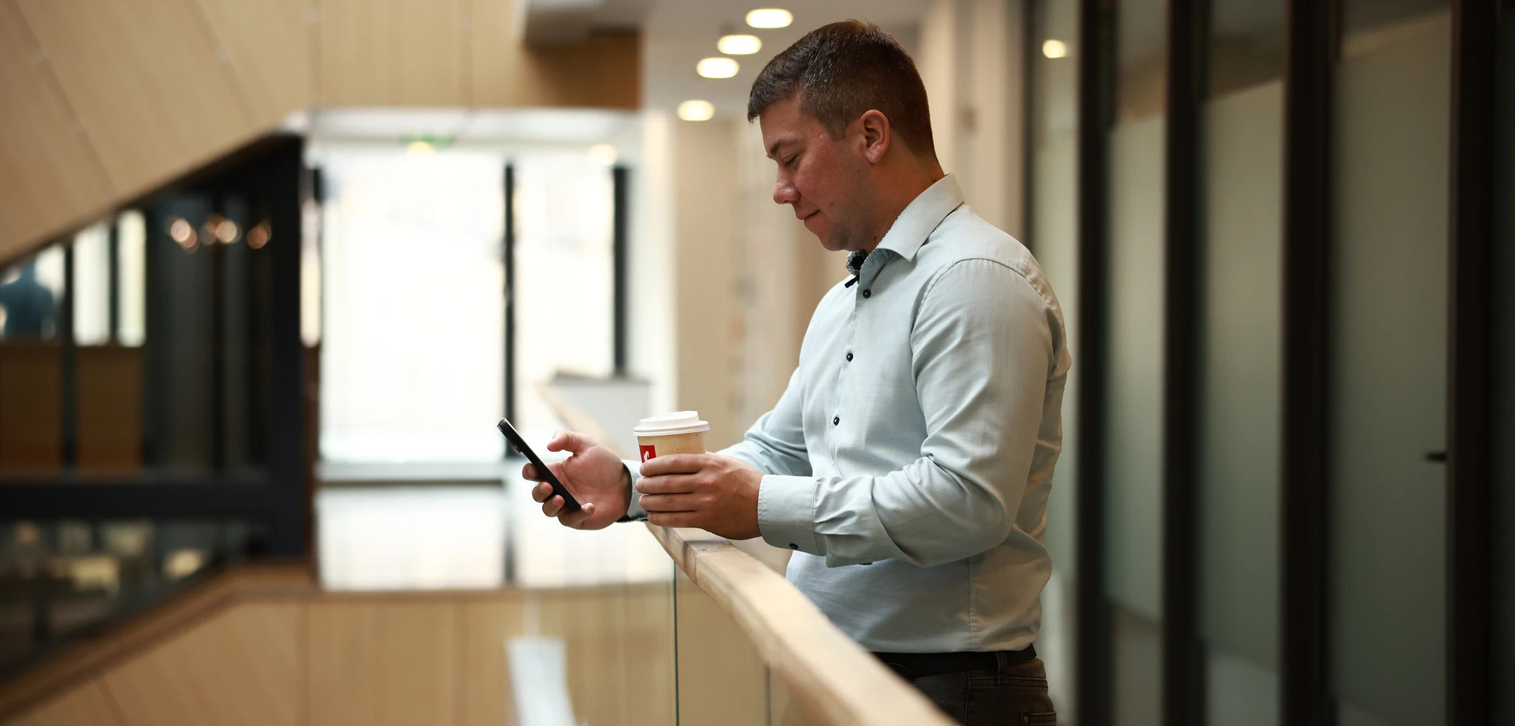 A man looking at his cell phone while holding a coffee cup. He is wearing a button-down shirt and is standing near a railing in a modern building.