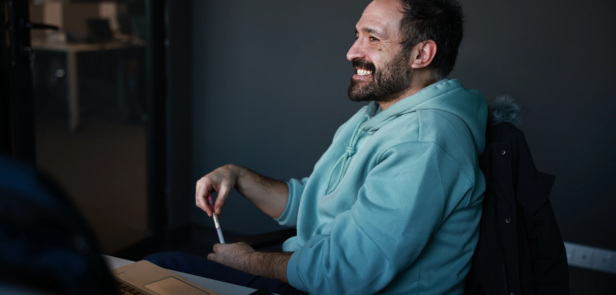 Smiling man with dark hair and beard, wearing a hoodie, holding a pen, in a modern office