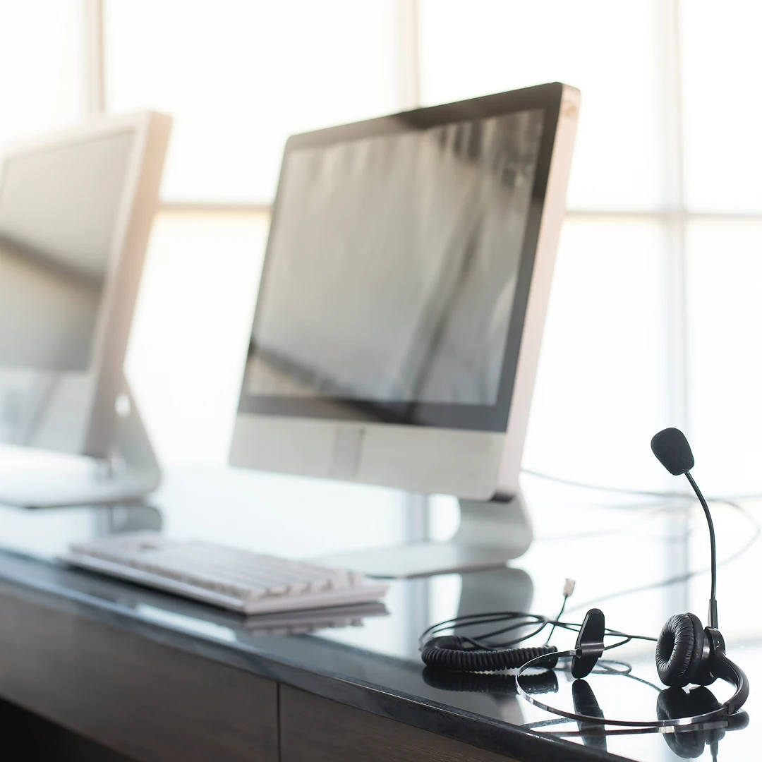 A black headset with a microphone rests on a black desk alongside a white keyboard and a desktop computer