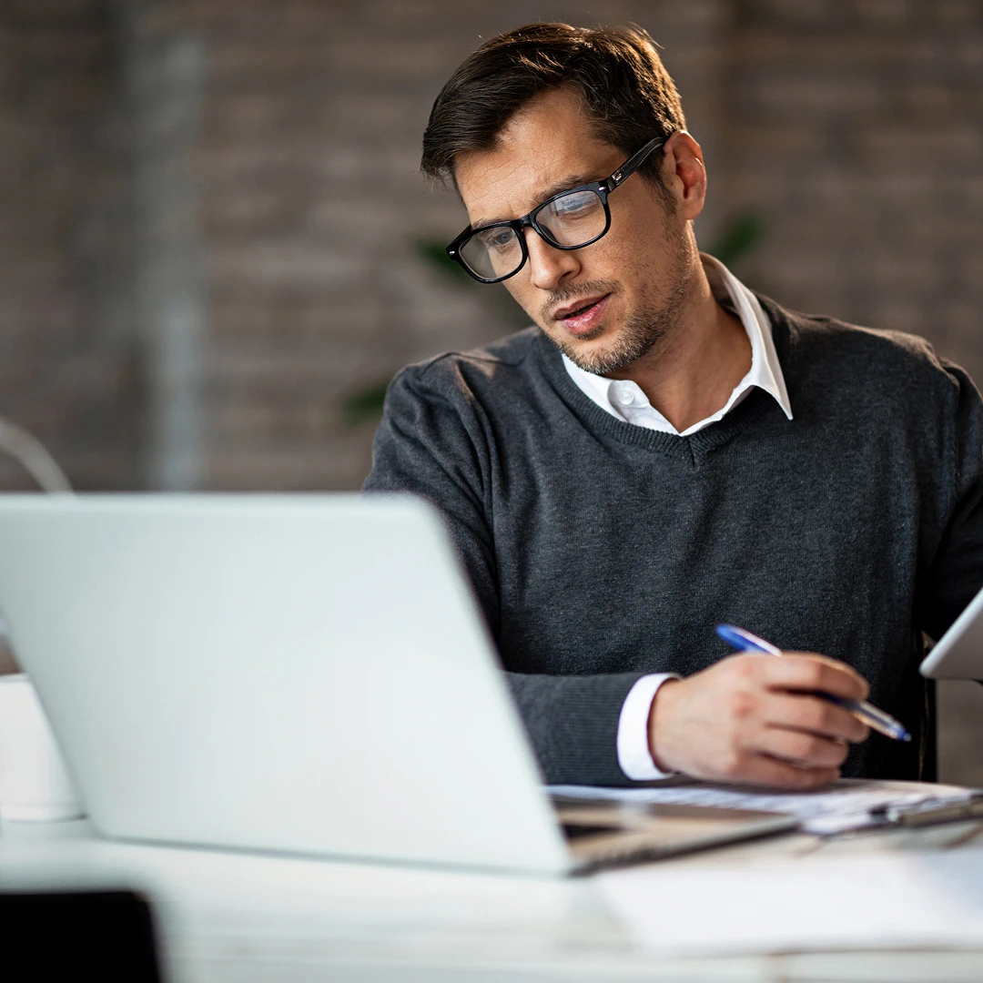A man with glasses and a black sweater is working on a laptop while holding a pen and paper