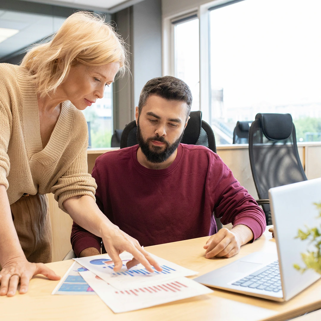 A blonde woman in a beige sweater pointing at graphs and charts on a desk, explaining data to a man in a burgundy sweater who is seated at the desk