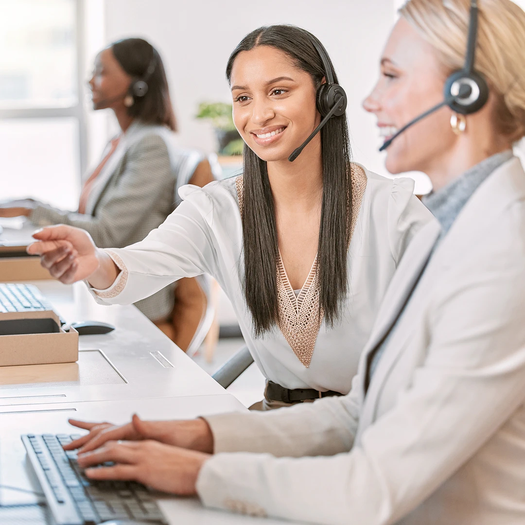 Two smiling women wearing white blazers and headsets, working together at a call center, with one pointing at the screen