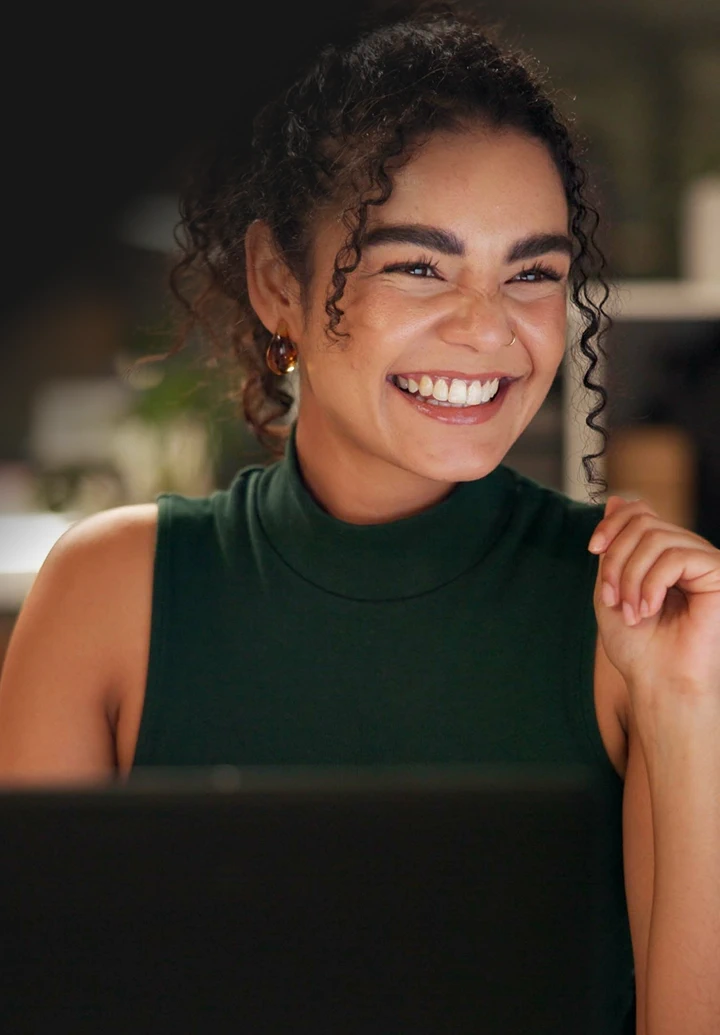 A smiling woman with curly hair, wearing a black t-shirt, is working on a laptop.