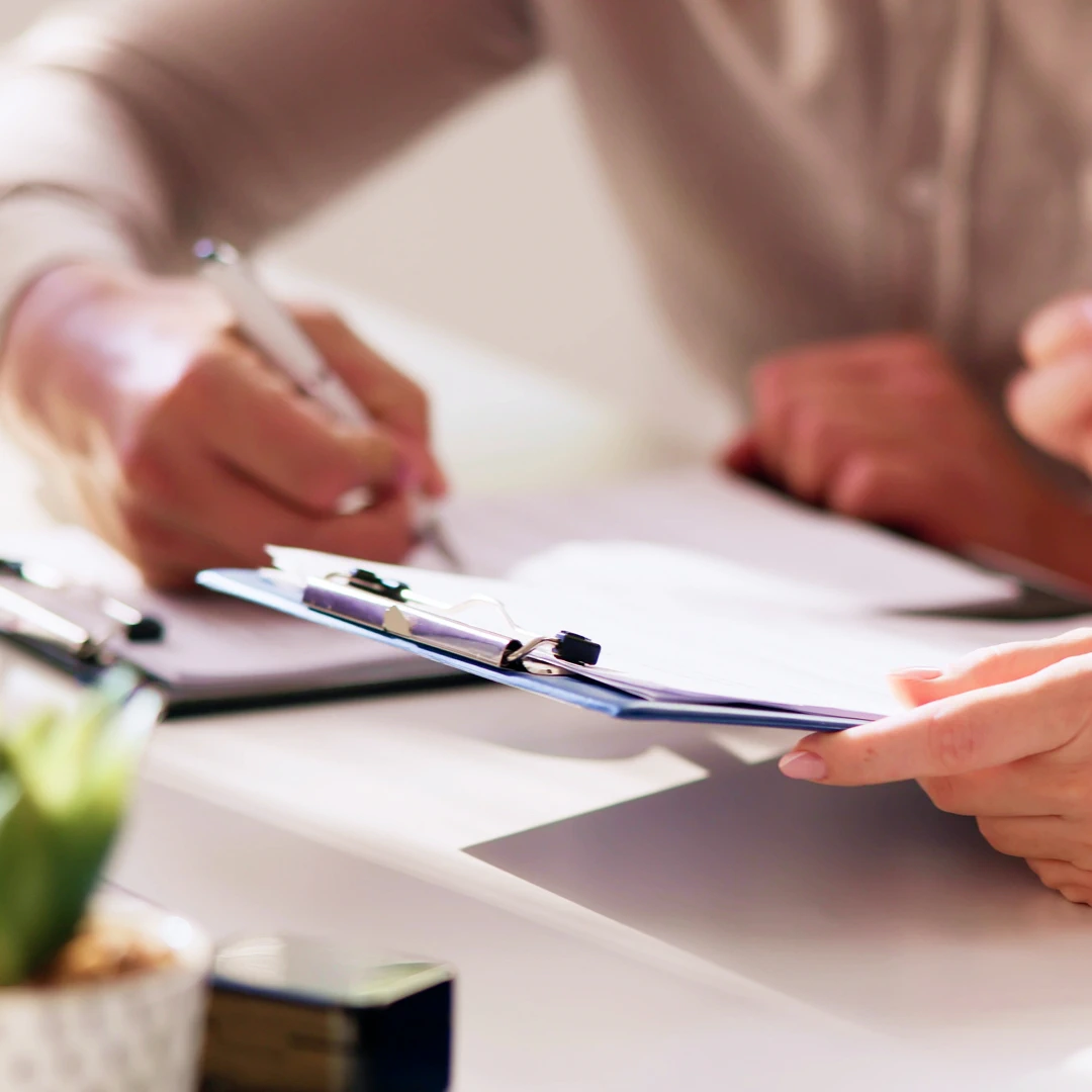 Close-up of a person's hands signing a document on a desk while another person holds a clipboard with papers