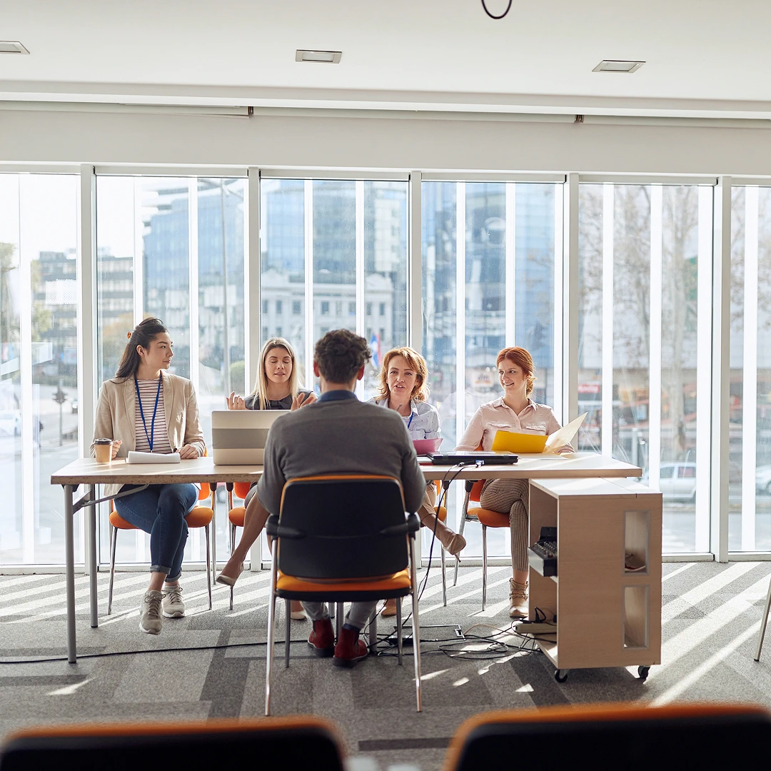 Four women seated at a table in front of a large window, facing a person sitting in a chair