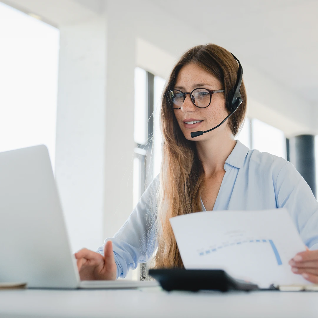 Female customer support agent with long brown hair, wearing a white blouse and a headset, reviewing data charts while working on a laptop