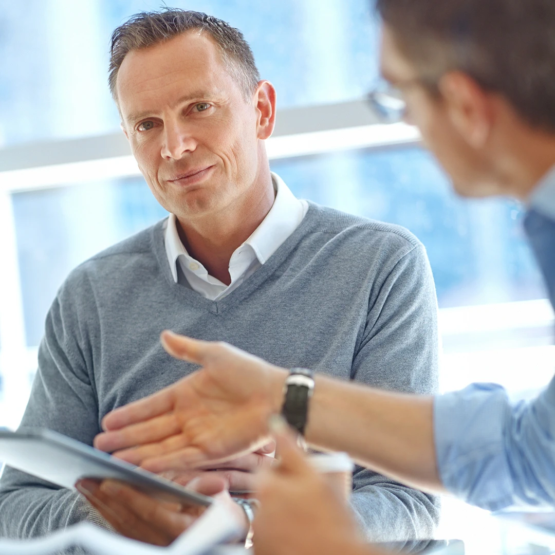 Man with short hair wearing a grey sweater listens intently while looking at a colleague pointing at a tablet