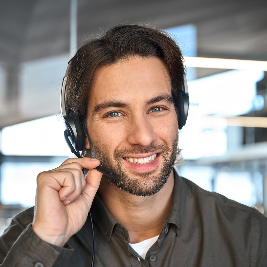Smiling customer support agent with brown wavy hair, blue eyes, and a stubble beard, wearing a dark grey shirt and a headset