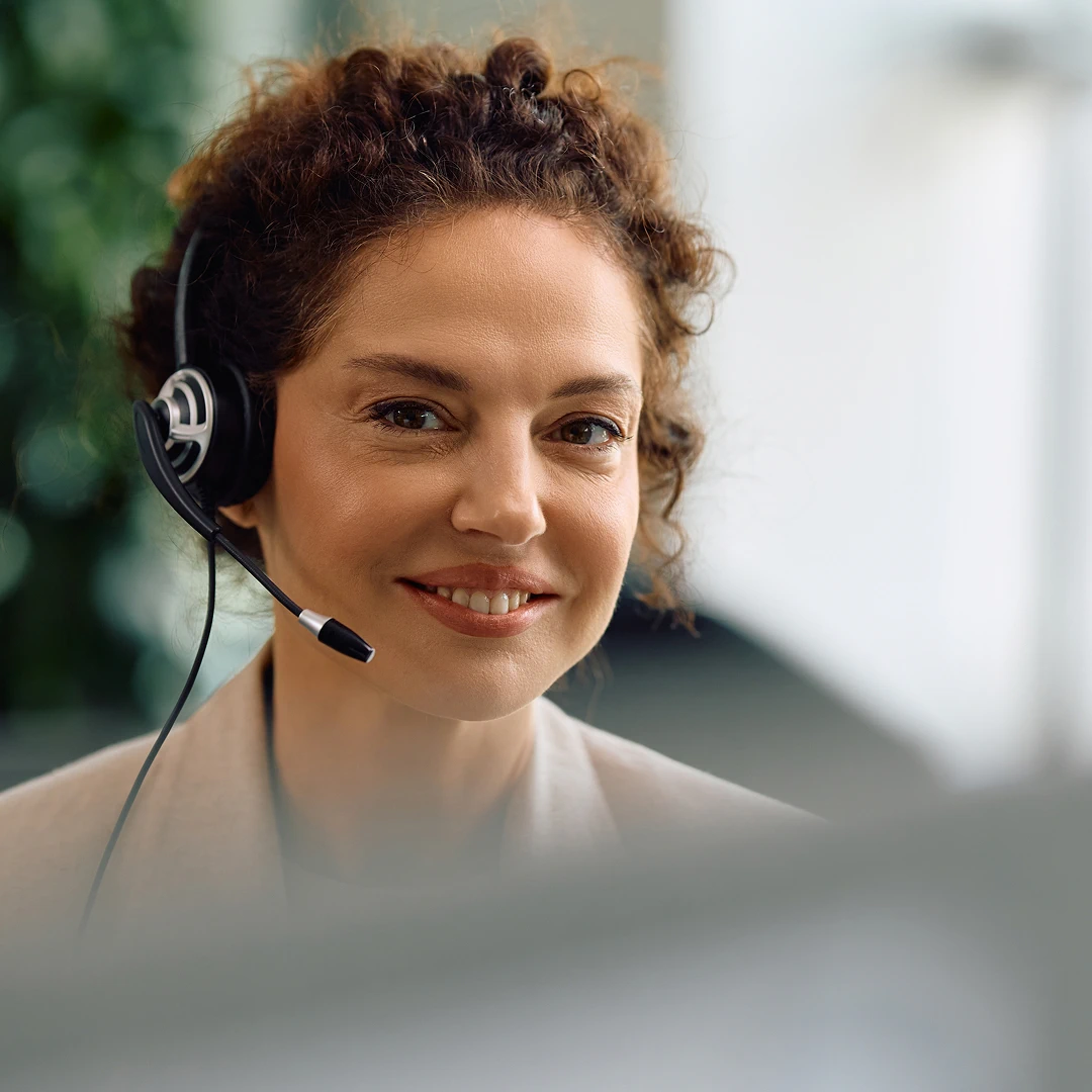 Friendly female customer support agent with curly brown hair, wearing a headset and a light-colored blazer