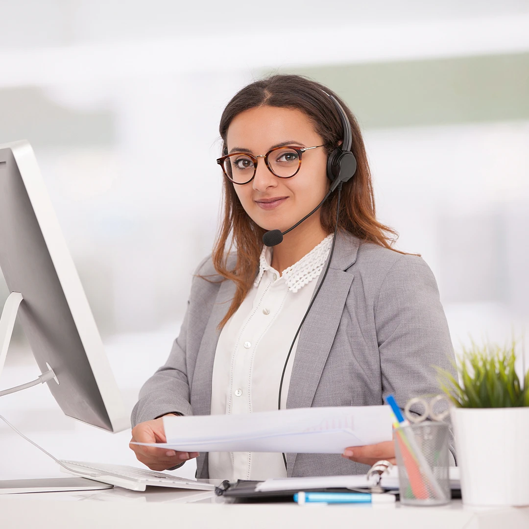 Focused female customer support agent with glasses and headset sitting at desk in front of computer