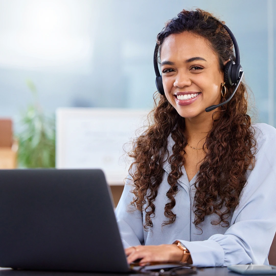 Smiling female customer support agent with long curly hair, wearing a headset, sitting in front of a laptop, and providing quality customer support