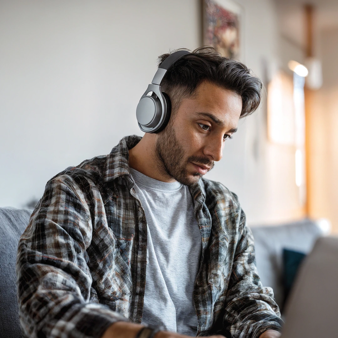 Latin American call center agent in a plaid shirt and headset, working on a laptop while sitting on a sofa
