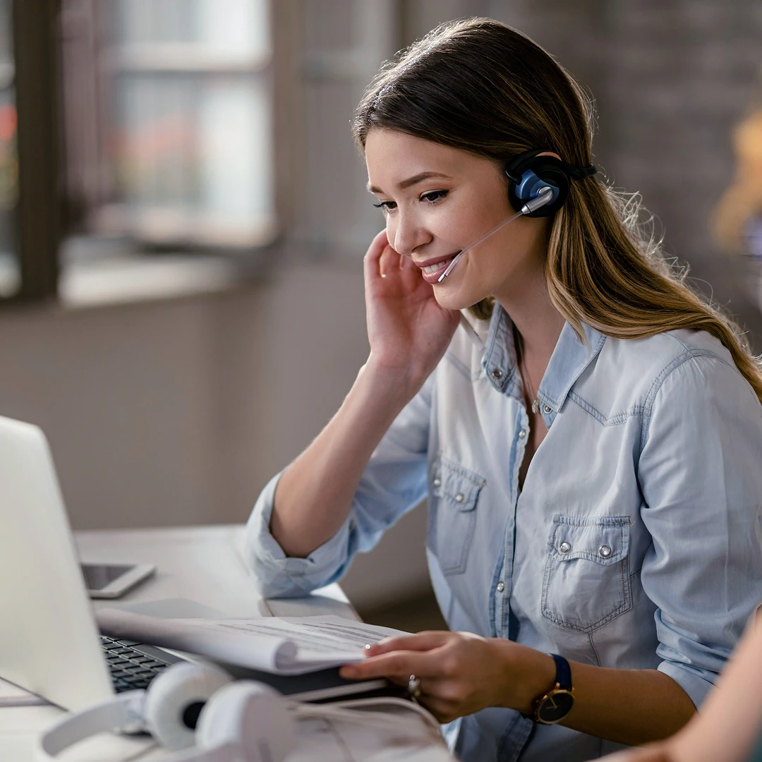 A young female customer support representative in a blue denim shirt, wearing a headset, working at a laptop