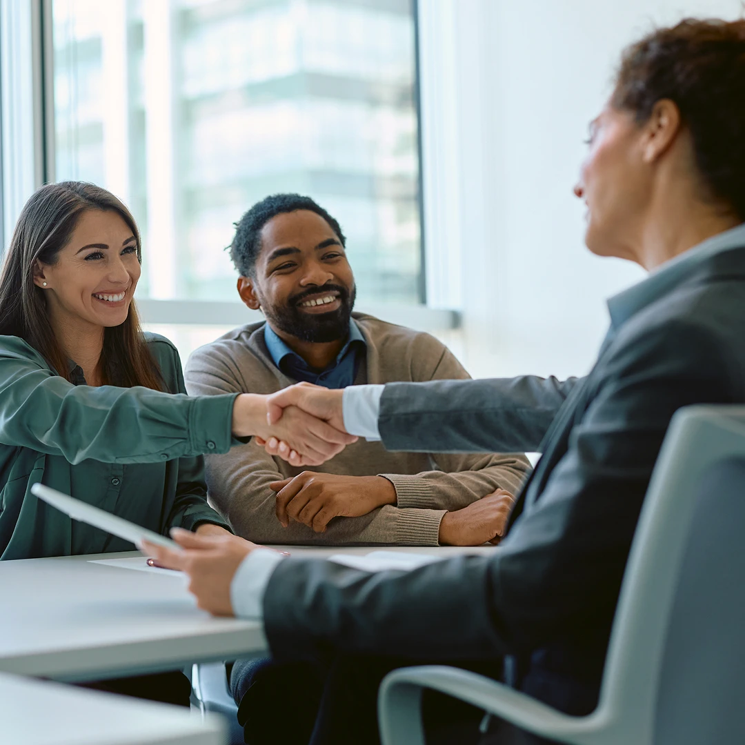 A business handshake taking place between two women while a smiling man sits beside them at a conference table