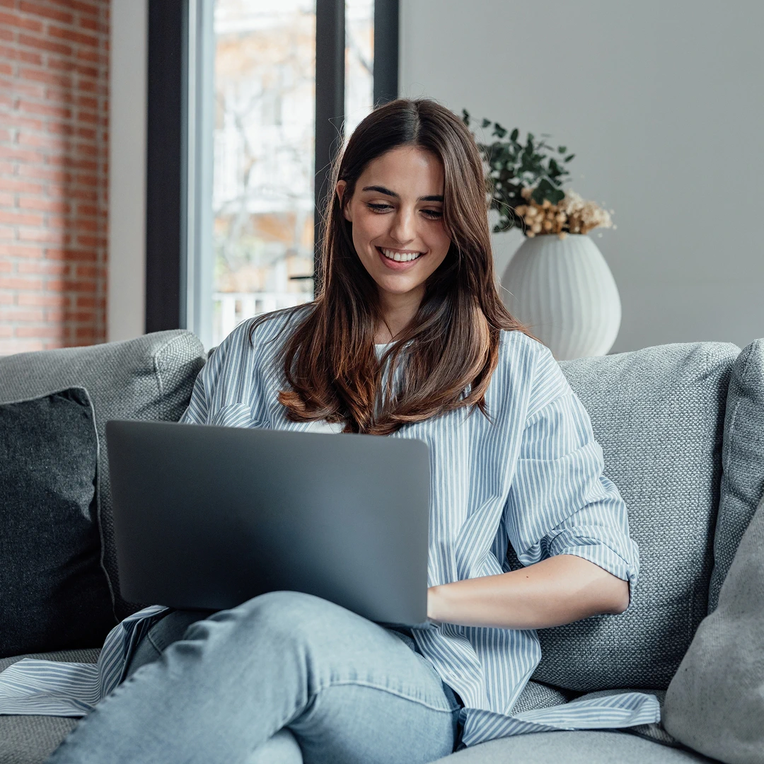 A young, pretty woman is working on a laptop while sitting on a comfortable gray sofa