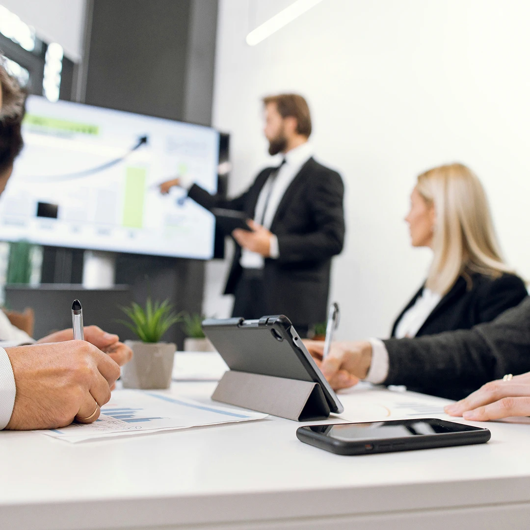 Team members in a business meeting with a presentation on a screen, taking notes and using a tablet