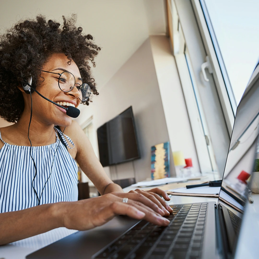 Smiling woman with curly black hair wearing a striped blouse and headset, working at a laptop