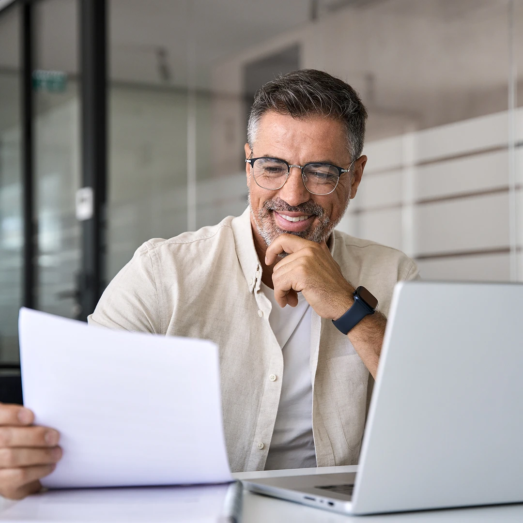 A smiling middle-aged man with glasses sits at a desk, reviewing a paper document while working on his laptop