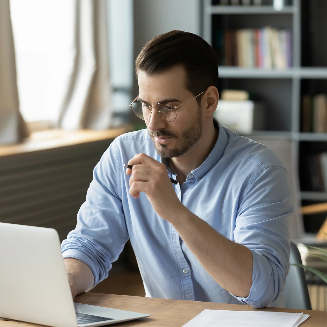 Man wearing glasses and a blue shirt, looking at a laptop and holding a pen
