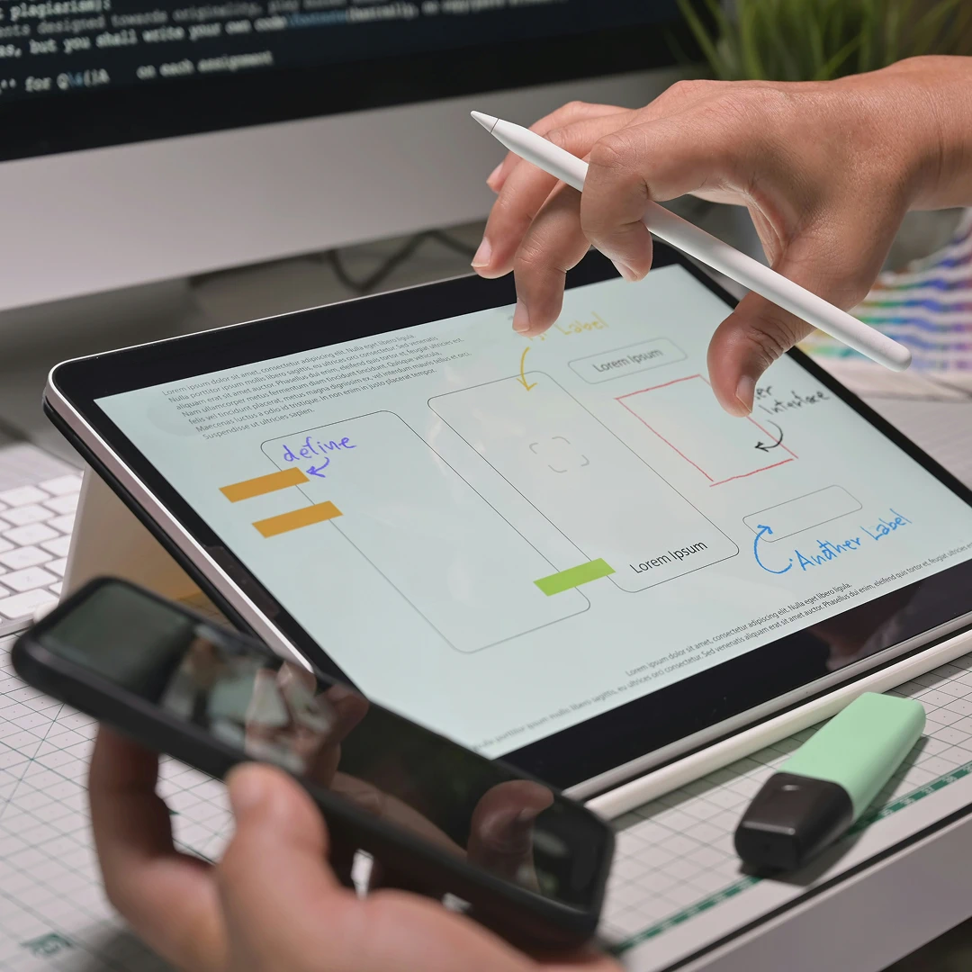 Hands holding a stylus and a smartphone while working on a tablet