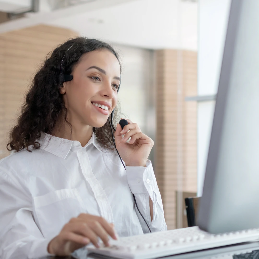 A smiling woman with curly hair, wearing a headset while working on a computer and providing customer support