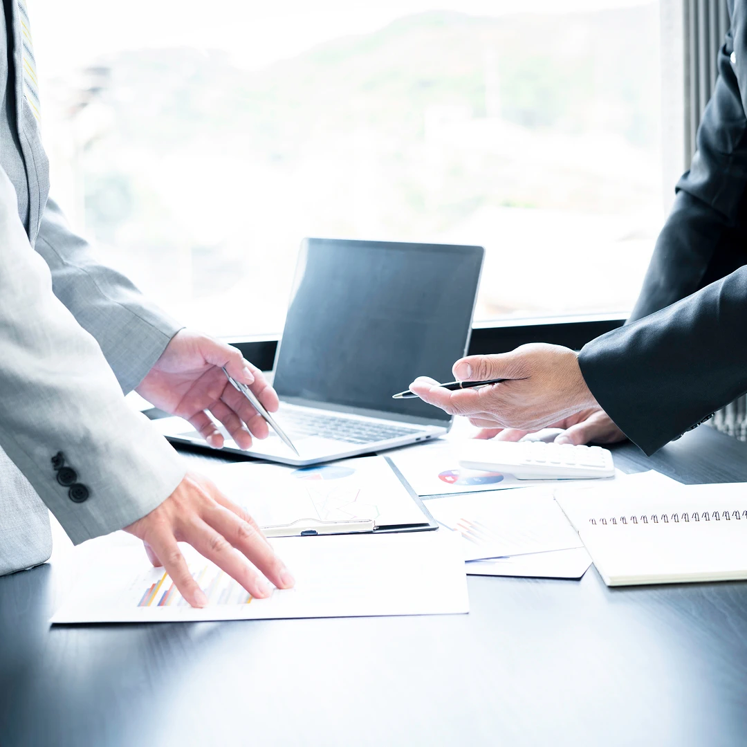 Two business professionals in suits discussing documents and data charts on a table with a laptop in a bright office setting
