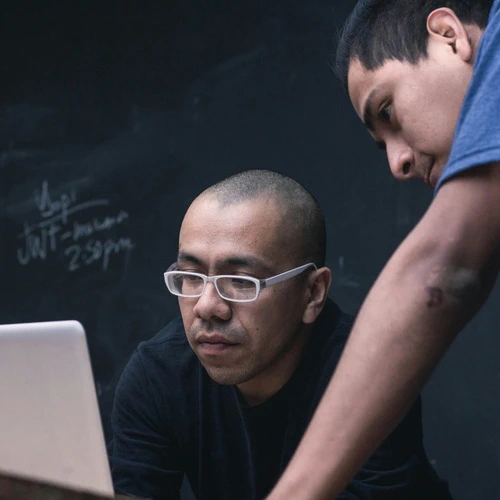 two asian men (one wearing white glasses) looking at a laptop screen, something is written on a blackboard behind them (only 2:50 PM is legible)