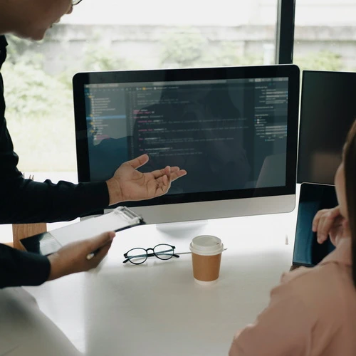 a man pointing to a computer screen with lines of code, a female looking at the screen, probably smiling