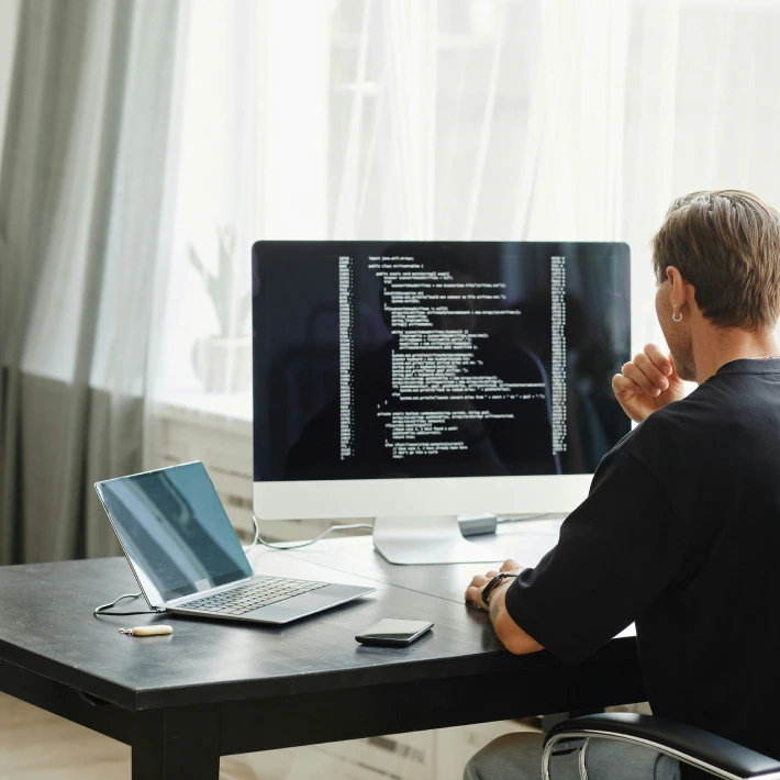 a man wearing an earring staring at a laptop connected to an Apple monitor, the monitor shows many lines of code. Apparently the man is a software developer
