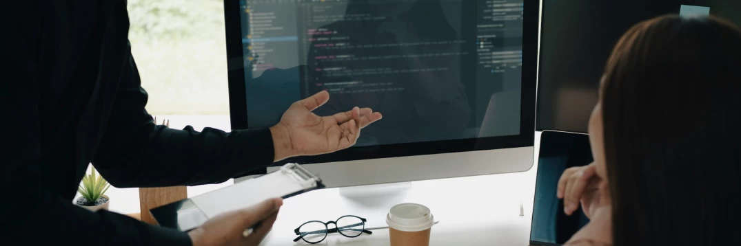 a man pointing to a computer screen with lines of code, a female looking at the screen, probably smiling
