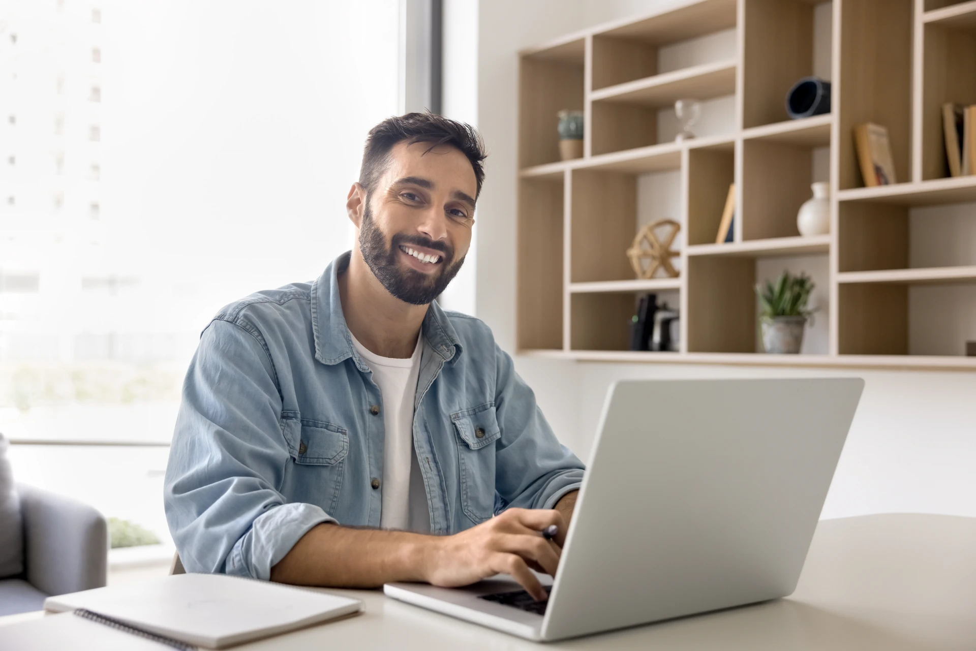 A smiling man with a beard, wearing a denim shirt, sits at a desk in front of a laptop