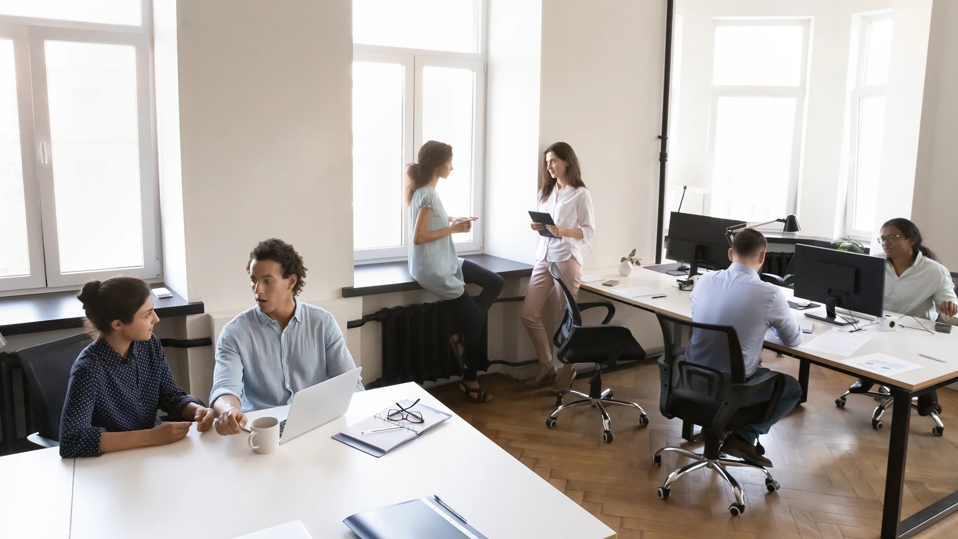 A group of people in a modern office setting, some working at their desks and others engaged in casual conversation near a window
