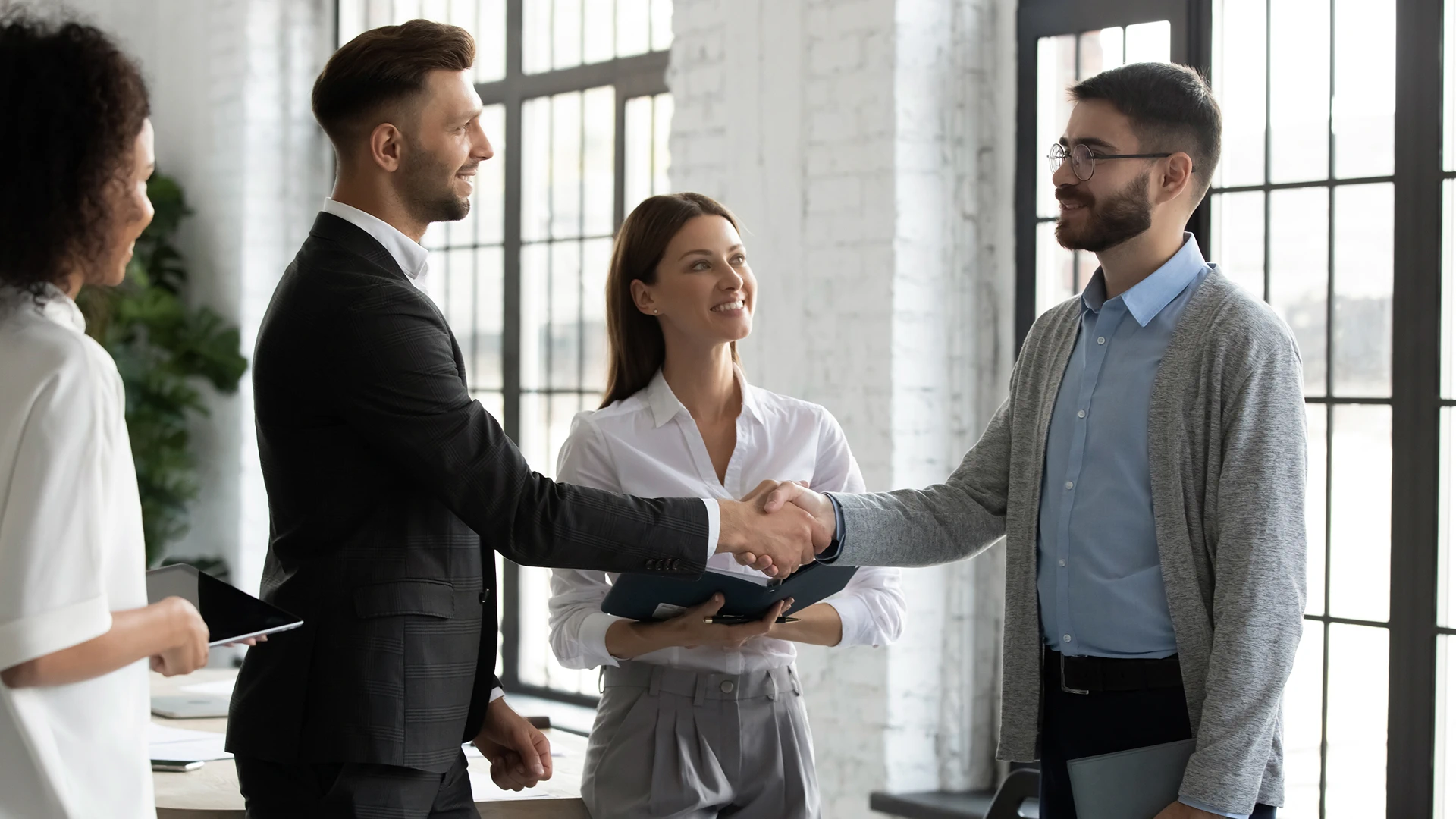 Two men are shaking hands, sealing an agreement, in front of two women in an office