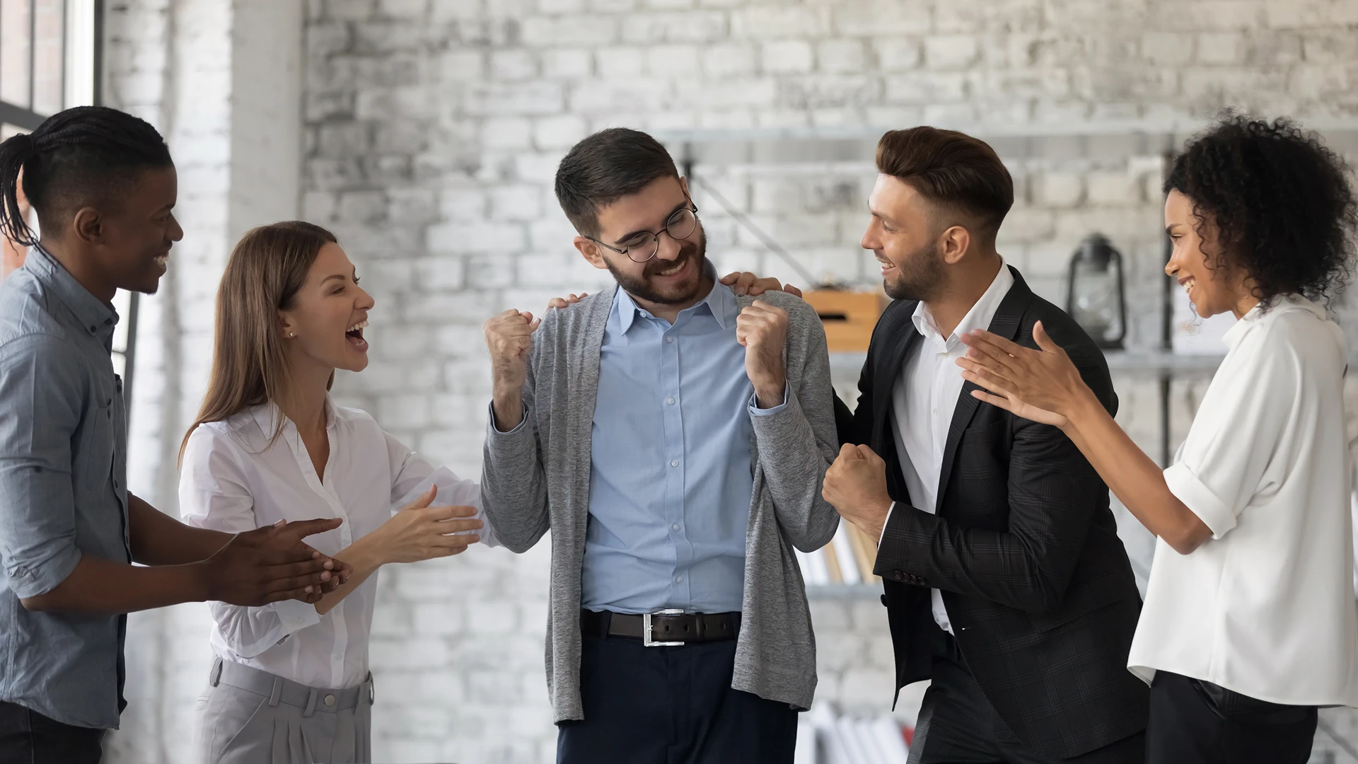 A group of colleagues is celebrating a man in a blue shirt and grey cardigan